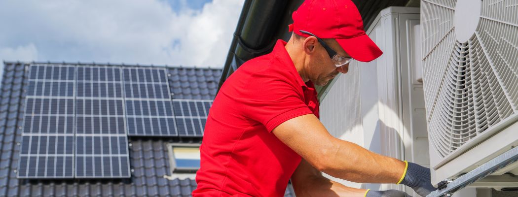 Mann mit rotem T-Shirt und Cap installiert W&auml;rmepumpe. Im Hintergrund ein Dach mit Solarmodulen